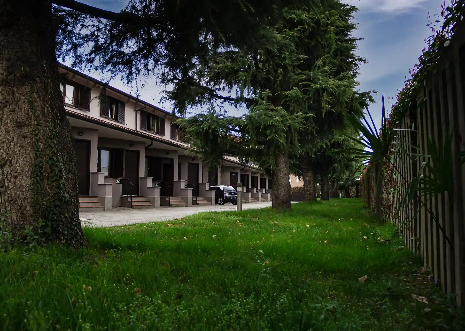 Nell'immagine si vede l'esterno dell'hotel con il prato e dei grandi alberi in primo piano. A destra c'è la recinzione. A sinistra c'è l'hotel con le stanze con ingresso indipendente e parcheggio dedicato accanto ad ognuno di essi.