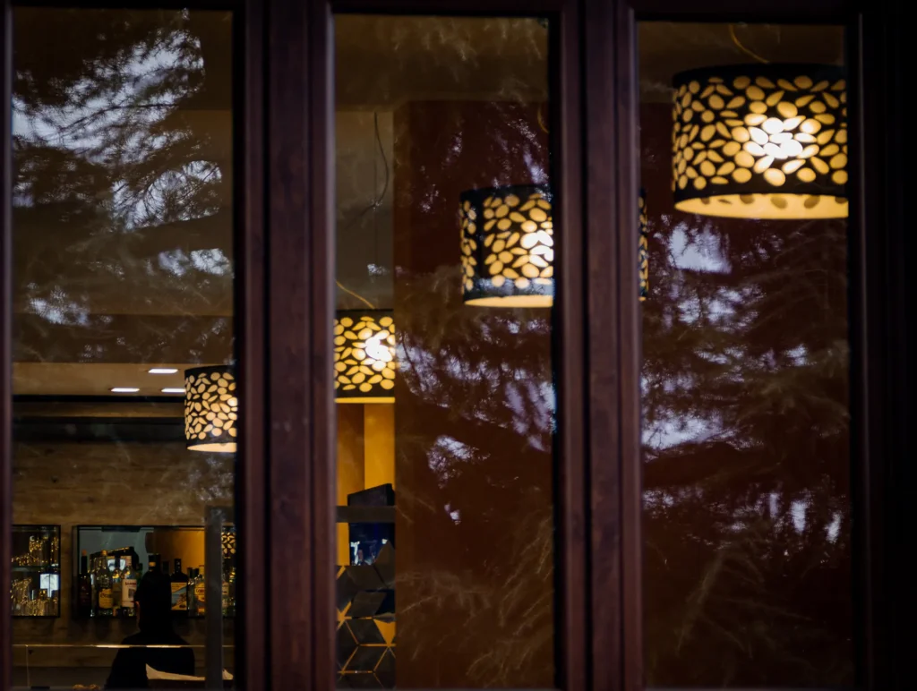 Nell'immagine si vede un dettaglio dell'hotel. Dall'esterno la prospettiva guarda attraverso la finestra dove si vedono 4 lampadari cilindrici neri ed oro e il bar sullo sfondo.