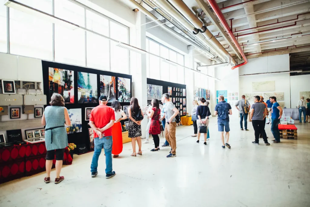 The image shows a warehouse where an art exhibition has been set up. There are various paintings and photographs hanging on the walls and supports and many people walking around admiring the works.